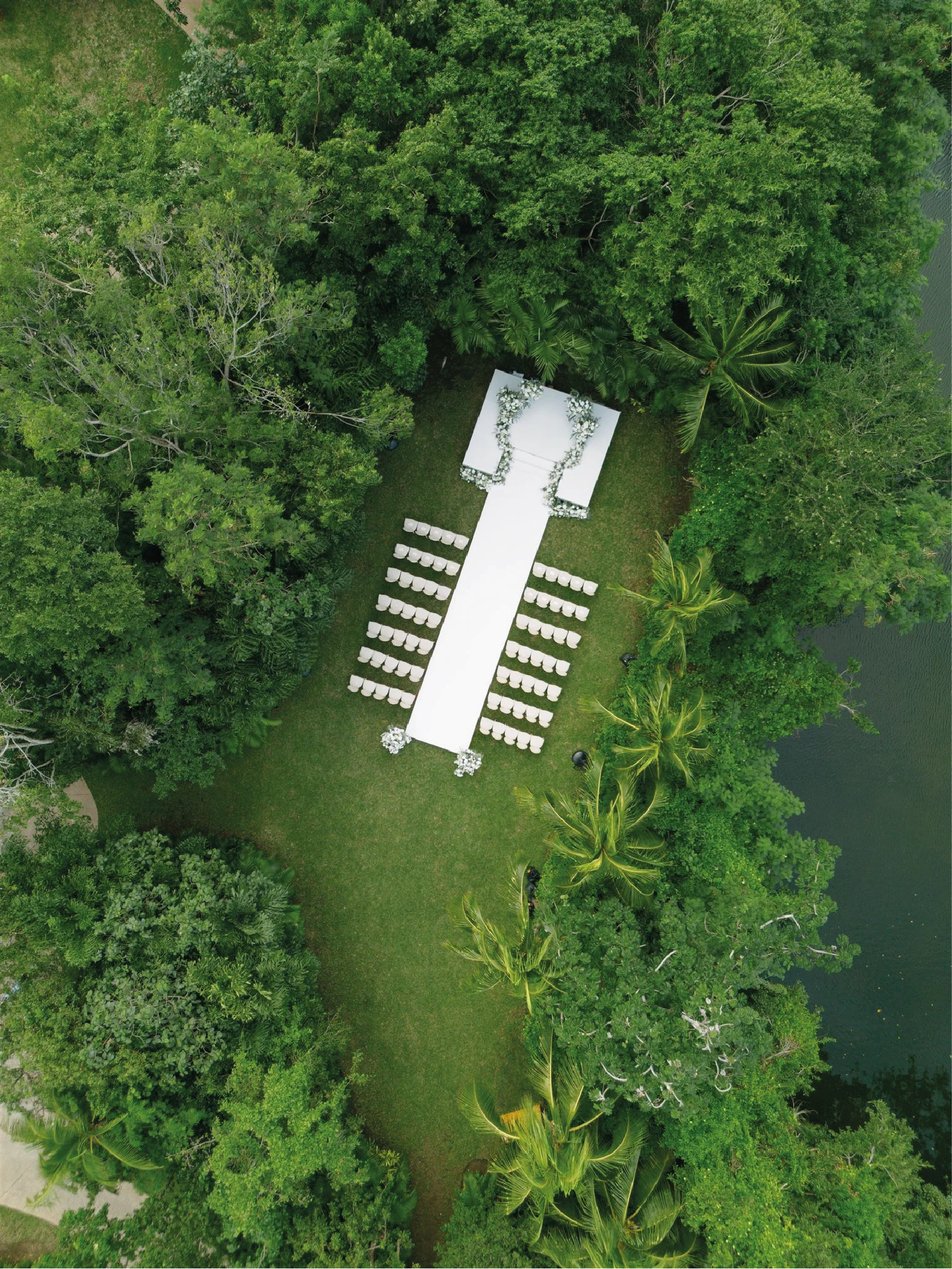 A stunning perspective of the Lagoon Lawn, a premier Rosewood Mayakoba wedding venue, featuring symmetric rows of white Louis XVI-style chairs lining a central white runway. The path leads to a large white floral altar with massive installations, set against a backdrop of dense tropical mangroves and the water's edge. This sophisticated setup for the wedding of Sam & Matt in Cancun Riviera Maya was designed and produced by Bash House.