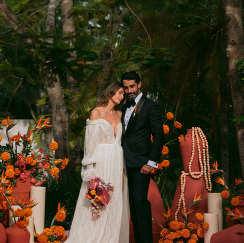 Alt Text: An elegant couple—she in a white dress and he in a black suit—at a tropical outdoor wedding reception at Belmond Maroma. The setting features striking Birds of Paradise floral decor with design and production by Bash House, illustrating the sophisticated aesthetic potential of premier destination wedding venues in Mexico.