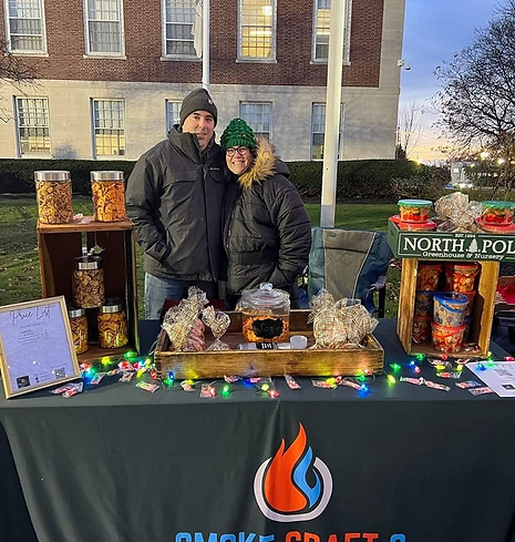 Two people bundled in winter coats and knit hats stand behind a SmokeCraft Q–branded table outdoors in front of a brick build