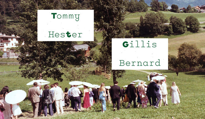 Two white placecards on cream textured paper displaying 'Tommy Hester' and 'Gillis Bernard' in green typewriter-style font, with the letters G and T highlighted in darker green. Cards are photographed against a blurred background showing wedding guests gathering on a grassy hillside in the Italian Dolomites with mountains and traditional alpine buildings visible.