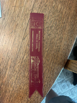 a maroon bookmark with gold writing stating "National Baseball Hall of Fame Cooperstown, New York", the Hall of Fame Logo, and the Hall of Fame Building