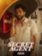 Man in white shirt uses a red payphone under a yellow hood. Background features black-and-white posters. Text: "The Secret Agent" and "NEON".