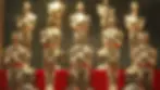Gold award statues in rows on a red surface, reflecting light, with a dark blurred background. Bright, shiny appearance conveys prestige.