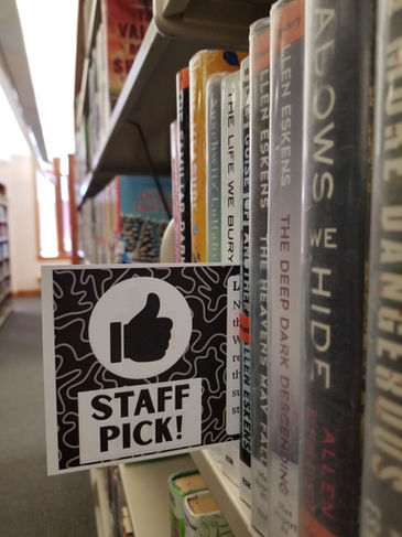 Staff pick insert tucked in between books on the library shelf. Insert features a black background with the white outline of organic shapes with a white circle with a thumbs up symbol and "Staff Pick"