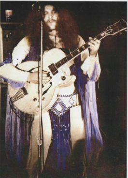 Musician with long hair and a fringe outfit plays an electric guitar on stage. Dim lighting creates a vintage, retro atmosphere.