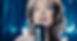A woman sings into a microphone on stage with a sparkling blue curtain backdrop. She wears earrings and shows a joyful expression.