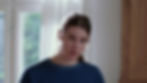 Woman in blue shirt looks surprised indoors, with natural light through a window. Simple wooden cabinet in the background.