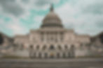 U.S. Capitol building with a cloudy sky, featuring grand white columns and a prominent dome. Steps and plants adorn the foreground.