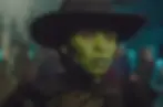 A person with green skin, wearing a black hat and dark attire, looks intently in a dimly lit setting with blurred figures in the background.