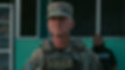 A man in camo uniform with "POLICE" vest stands seriously outdoors. Background has a teal and black building; another person is blurred.