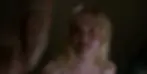 Blonde woman with a neutral expression in a dimly lit room, looking at a man out of focus on the left. She's wearing a sparkly top.