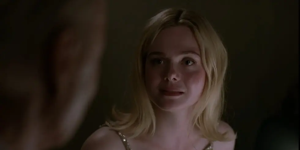 Blonde woman with a neutral expression in a dimly lit room, looking at a man out of focus on the left. She's wearing a sparkly top.