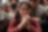 A woman in a red dress stands among a crowd, hands clasped in front of her face, looking distressed. The background is blurred.