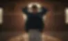 Man stands with hands behind head in a dim room with a circular window. Wears a dark shirt and jacket. Sunlight highlights the sandy floor.