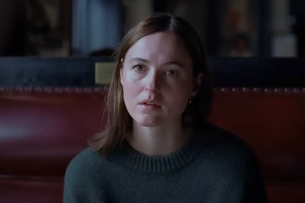 A woman with a thoughtful expression sits in a dimly lit room. She's wearing a dark sweater against a red leather seat, hinting at a somber mood.