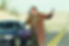 Man in plaid coat stands on deserted road, holding a shotgun and phone. Car nearby. Desert landscape, yellow lines on road.