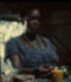 Woman in a plaid dress lights a candle in a dim room, wearing turquoise beads. Background shows a window with blurred foliage. Mood is solemn.