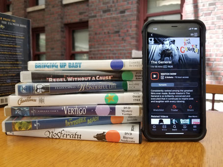 Stack of classic DVDs on a table next to a smartphone displaying "The General" film info. Background shows a brick wall and windows.