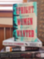 A group of books including Upright Women Wanted, Five Total Strangers, Hounded, and Written in Red stacked in a library chair.