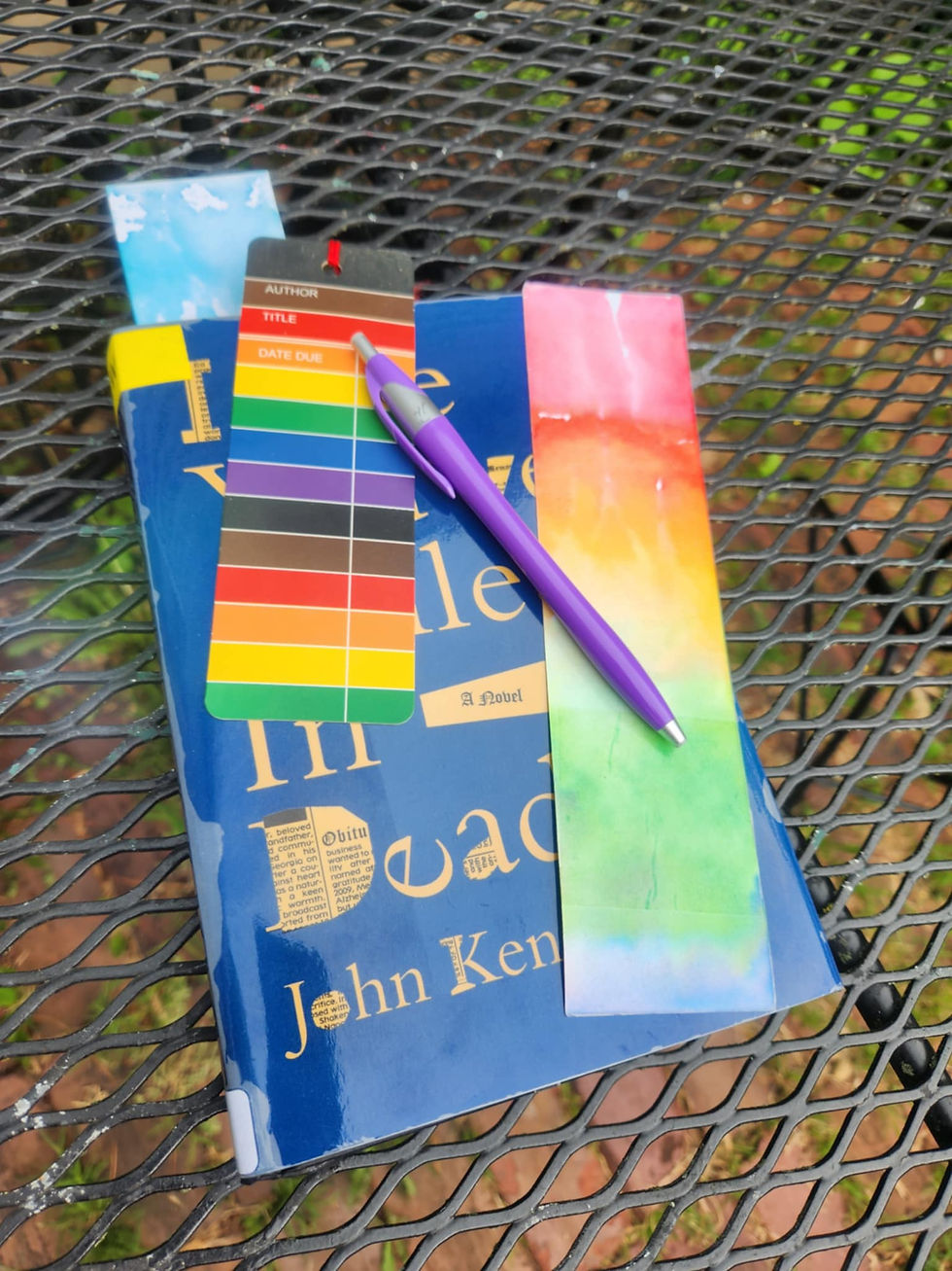Blue book on metal table with colorful bookmarks, purple pen, and outdoor greenery. Text on bookmarks includes "AUTHOR" and "TITLE".