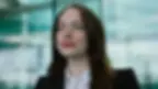 Woman smiling confidently in a business suit, with long brown hair, against a blurred modern glass building background.
