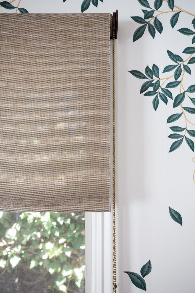 A beige woven window blind partially covers a sunlit window with a leafy pattern on white walls. A chain is used to adjust the blind.