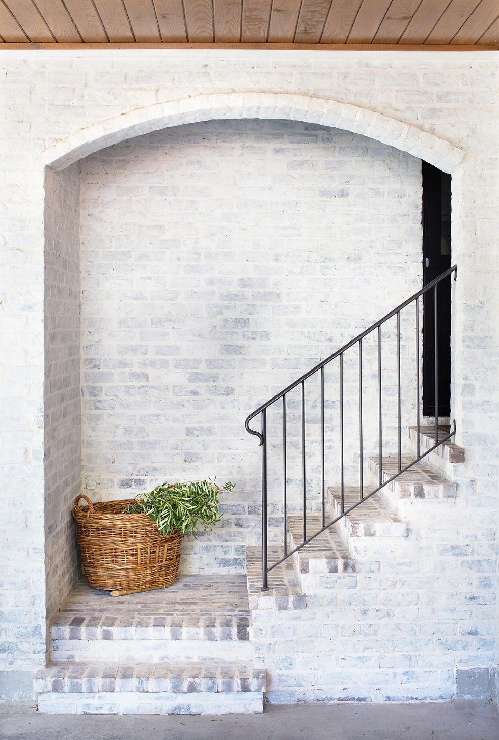 Wicker basket with greenery on brick steps under a white brick arch. Metal railing leads upstairs. Wooden ceiling adds warmth.