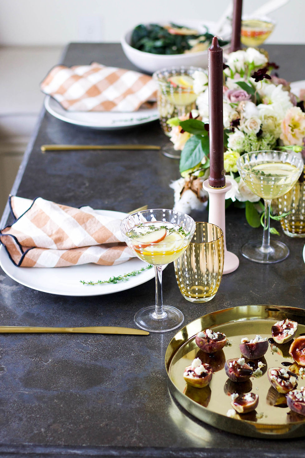 Elegant table setting with cocktails, figs with cheese, and floral centerpiece. Beige napkins, gold cutlery, and candle. Festive mood.