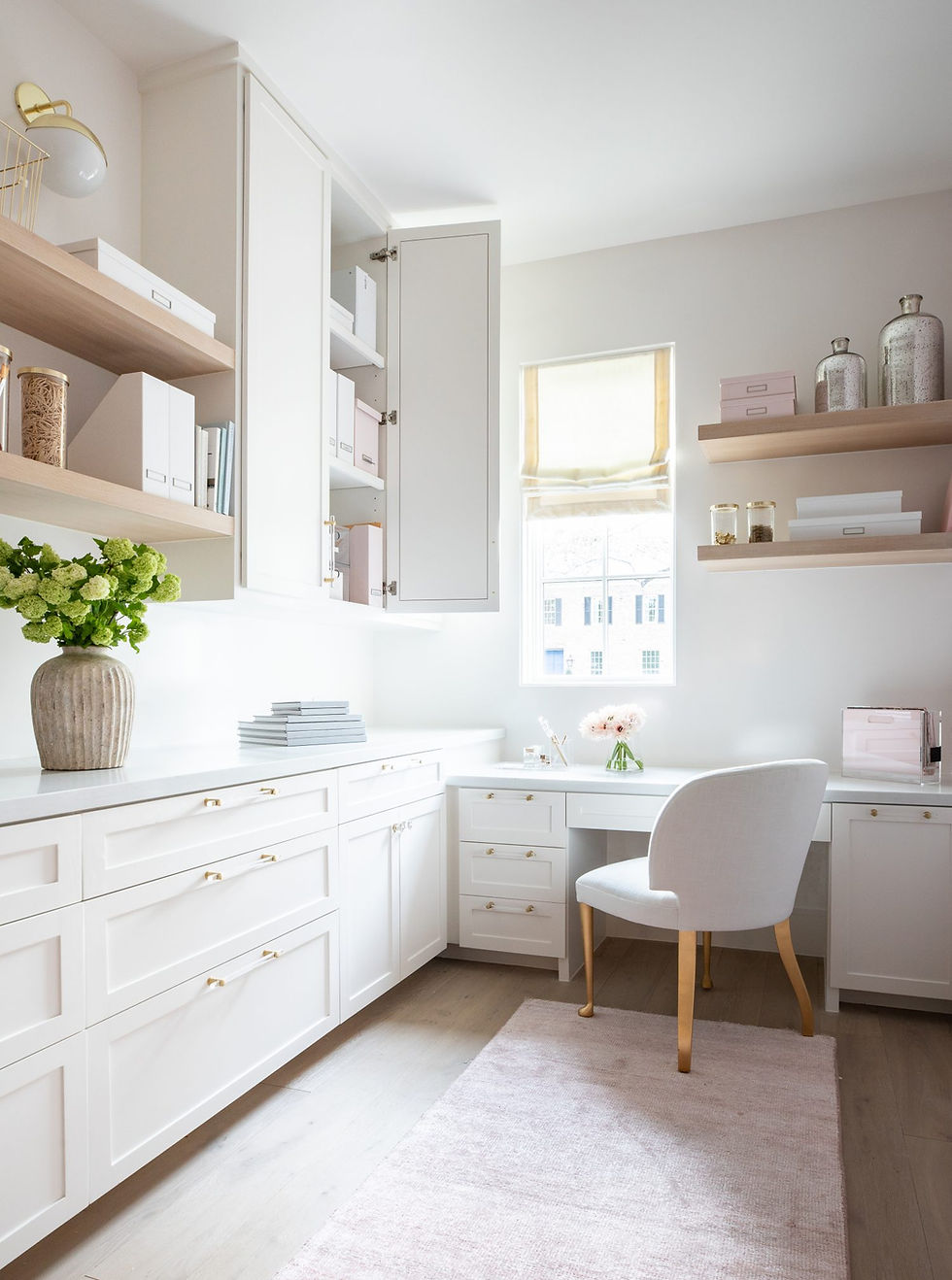 Bright home office with white cabinetry, shelves with boxes, a vase of flowers, and a soft pink rug. Sunlight streams through a window.