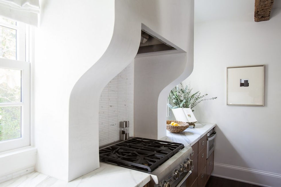 White kitchen with a curved range hood, gas stove, and marble counter. A basket with lemons and a plant beside a window. Calm, bright mood.