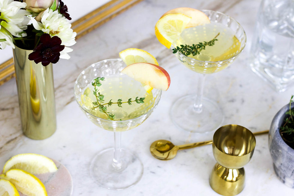 Two Autumn Fizz with lemon, apple slices, and thyme on a marble table. Gold jigger, floral vase, and lemon slices nearby.