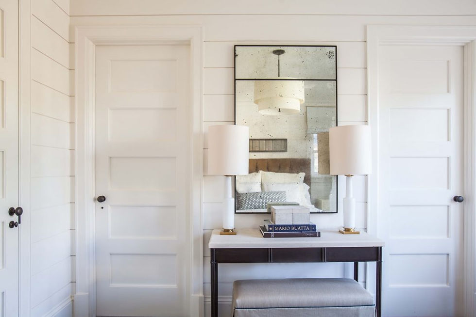 Elegant room with a large mirror reflecting a bed, flanked by white lamps on a console table. Books titled "MARIO BUATTA" sit below. Cozy vibe.