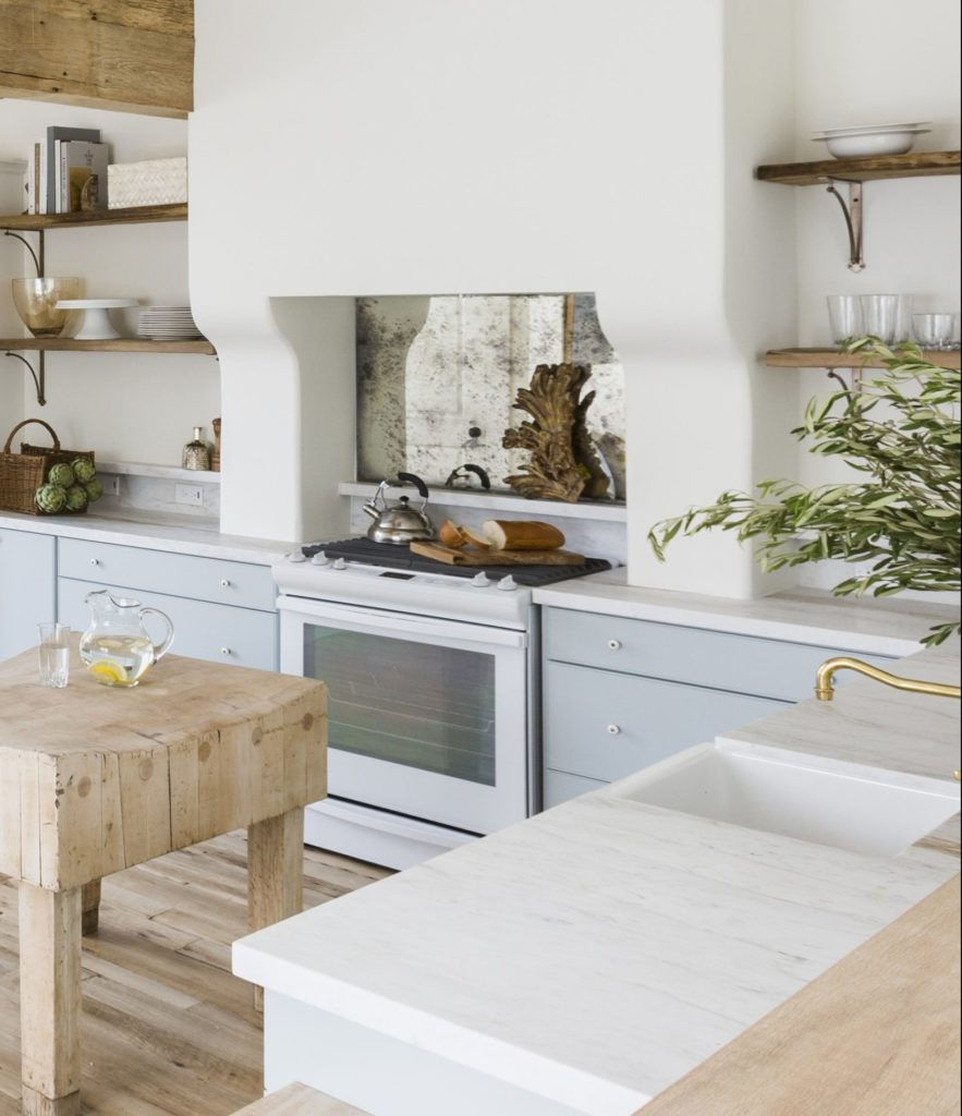 Rustic kitchen with light blue cabinets, wooden shelves, and a butcher block island. Lemon water on the counter, greenery adds a fresh touch.