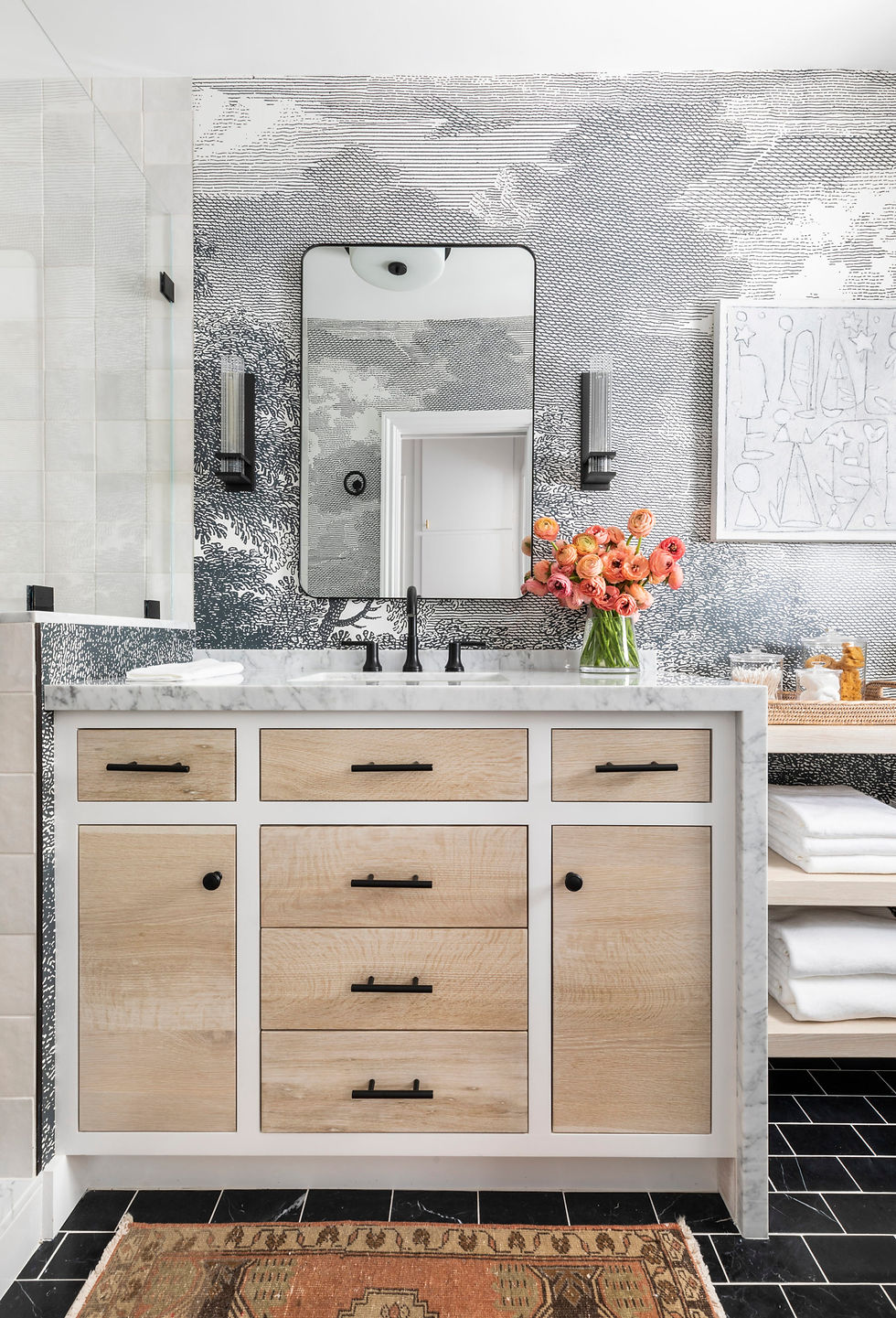 Modern bathroom with wood vanity, marble countertop, and black fixtures. Vase of orange flowers, abstract wall art, and patterned wallpaper.