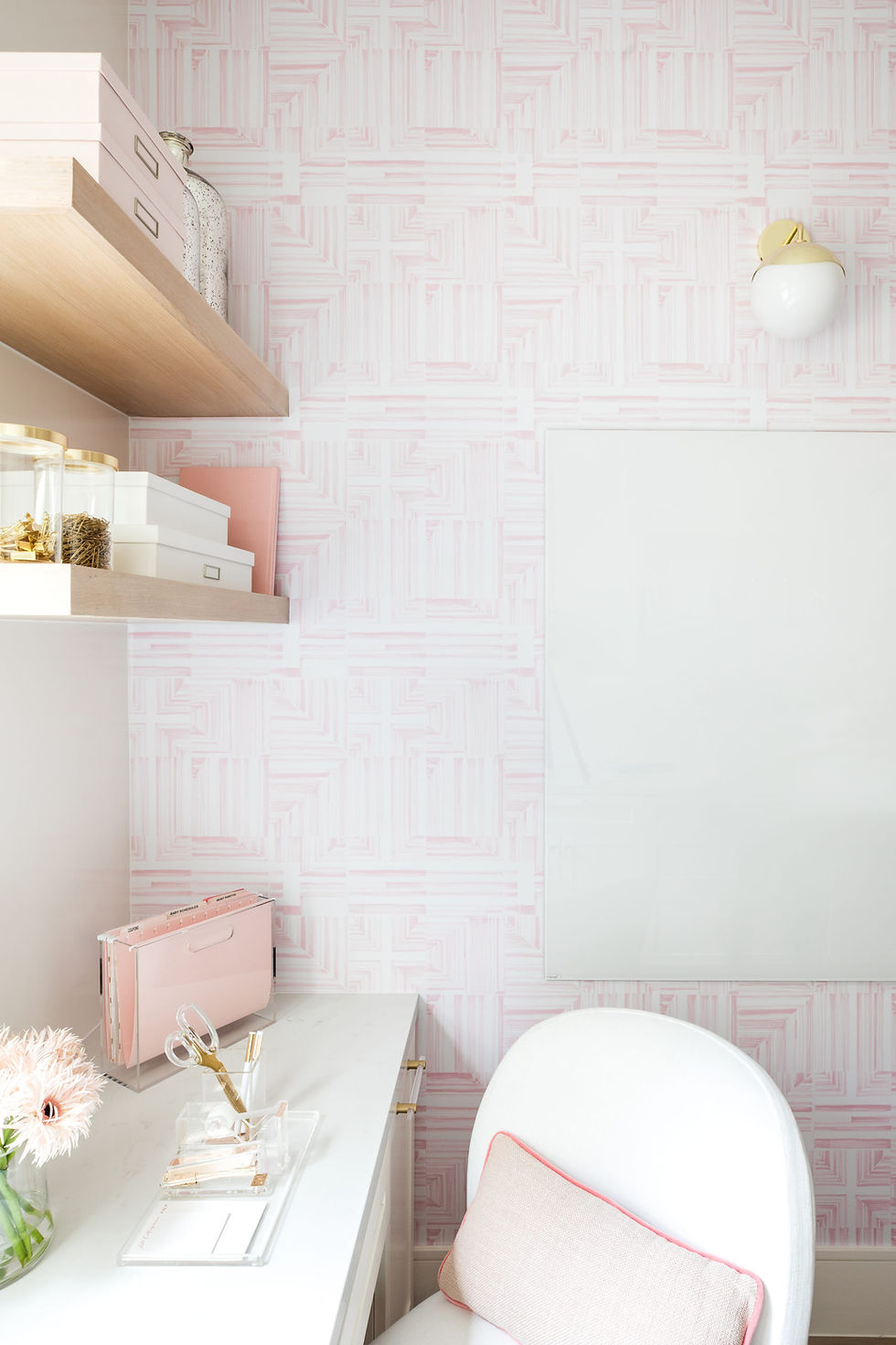 Stylish office with pink geometric wallpaper, white desk, and modern decor. Shelves hold boxes; flowers add a fresh touch. Cozy vibe.