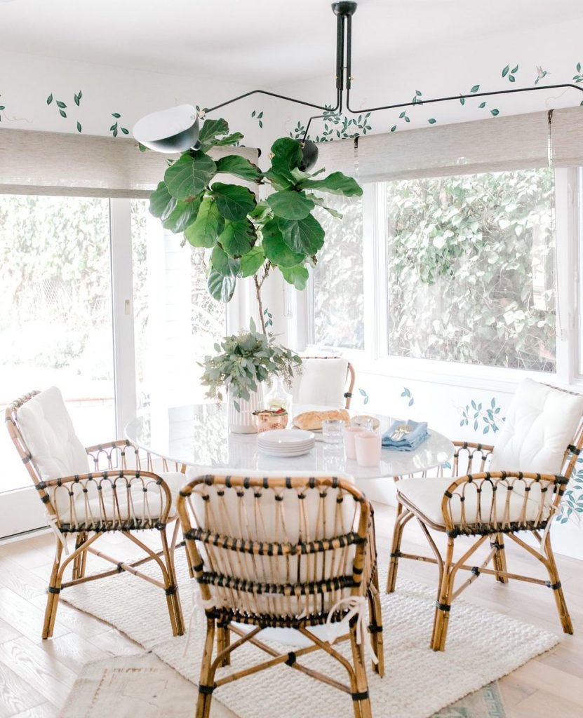 A bright dining area with a glass table, four wicker chairs, and a leafy plant. Plates, glasses, bread, and cloths are set. Leafy wall decor.
