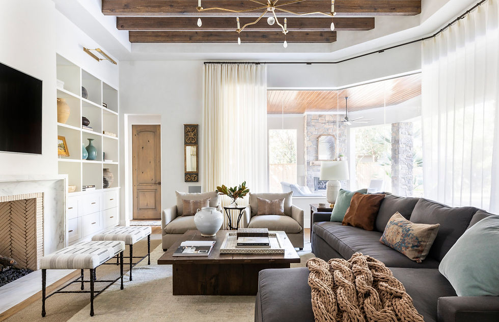 Cozy living room with beige sofas, dark coffee table, and TV. Wooden beams, white curtains, and shelves with decor. Natural light fills space.