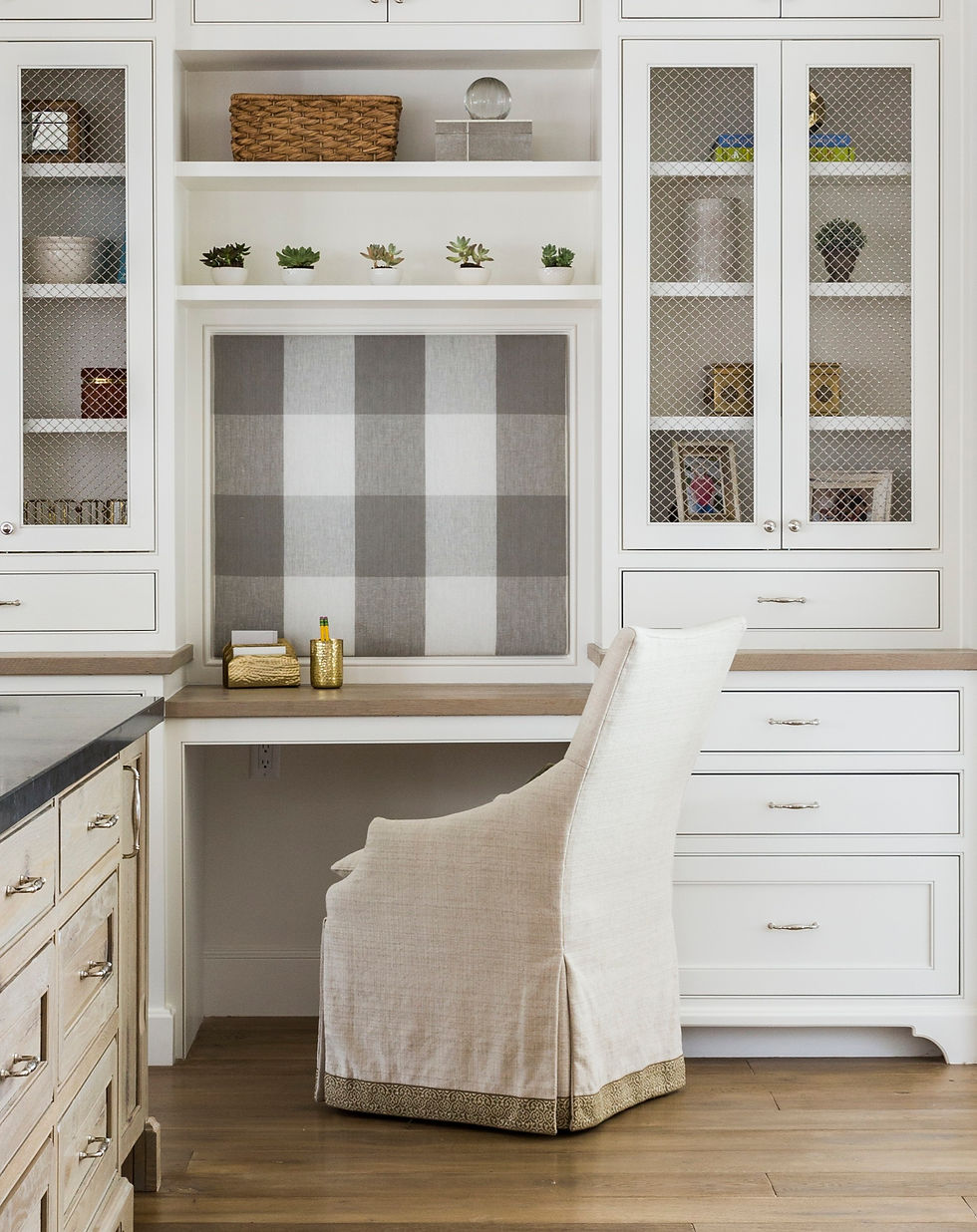 Cozy home office with white shelves, succulents, and a plaid accent wall. Beige chair in front of a wooden desk creates a calm setting.
