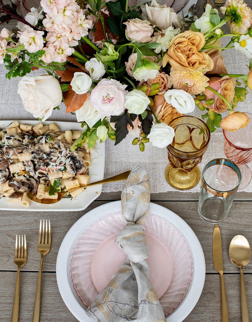 Elegant table setting with colorful flowers, rigatoni pasta dish, pink and white plates, gold utensils, and glassware on a wooden table.