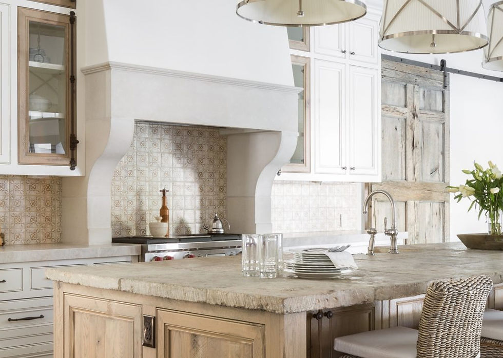 Rustic kitchen with a stone island, farmhouse sink, and wooden cabinets. Flowers on the counter, soft lighting, and tile backsplash.