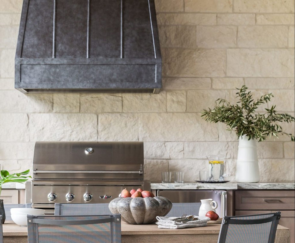 Outdoor kitchen with stainless grill against stone wall. Table with fruits, potted plant, and glass pitcher. Calm and inviting setting.