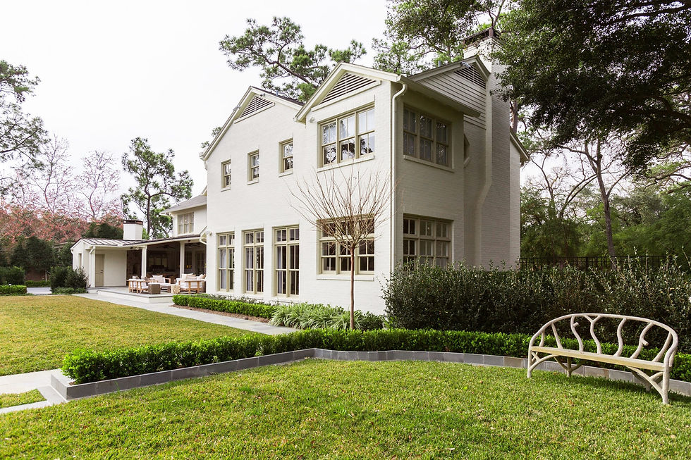 House with light bricks, large windows, and a patio furnished with chairs. Lawn and trees surround it. A bench sits on the grass. Tranquil setting.