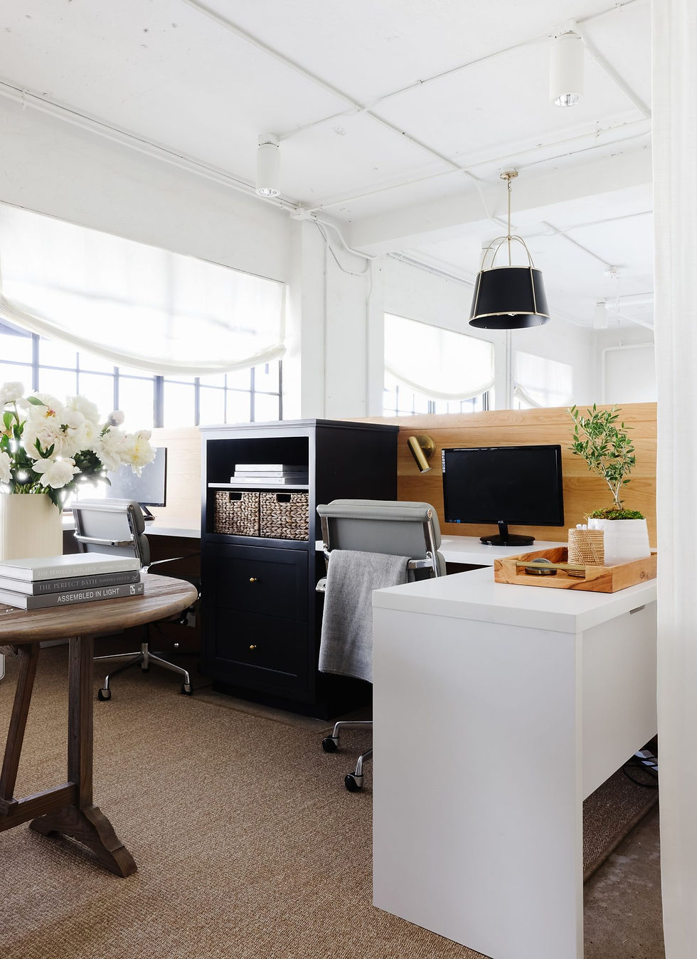 Modern office with white desks, chairs, monitors, a plant, books, and flowers. Neutral tones, large windows, and overhead lighting create a bright ambiance.