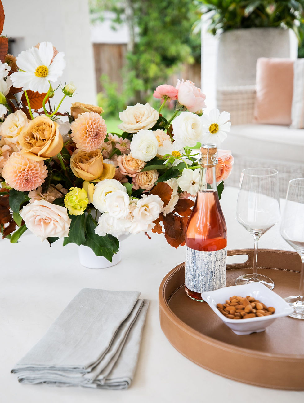 A bouquet of pastel flowers, a bottle of rosé, almonds, and wine glasses on a brown tray. Soft-pink pillows and greenery in the background.