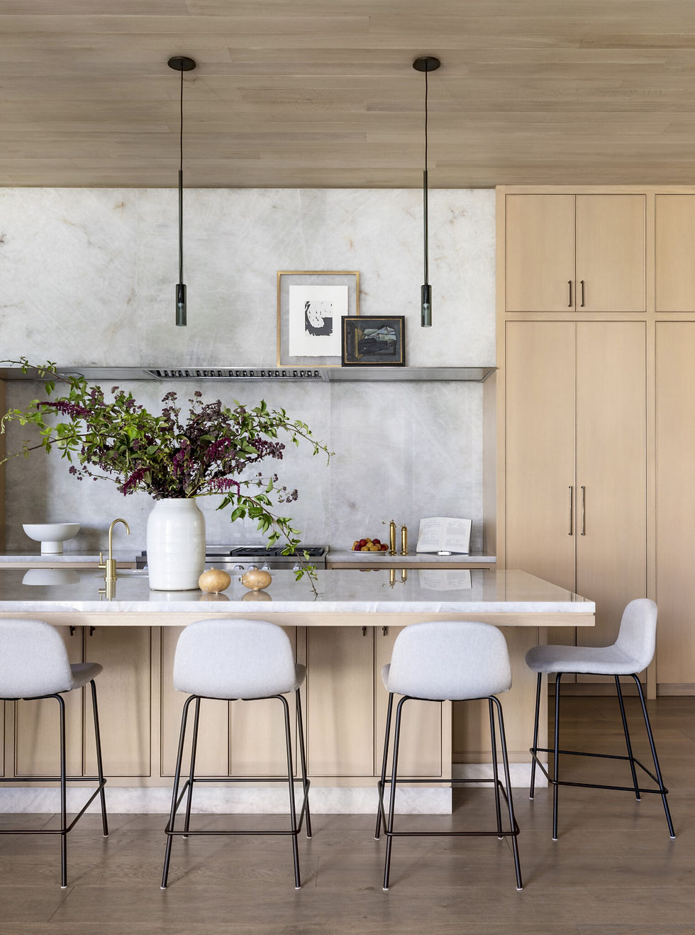Modern kitchen with a marble island, light wood cabinets, and gray stools. A vase of purple flowers and wall art add elegance.