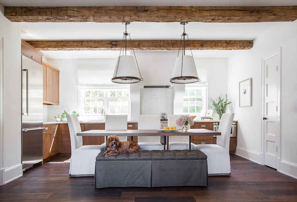 Kitchen with wooden beams, white chairs, and hanging lights. A brown dog rests on a gray bench. Neutral tones and bright windows.