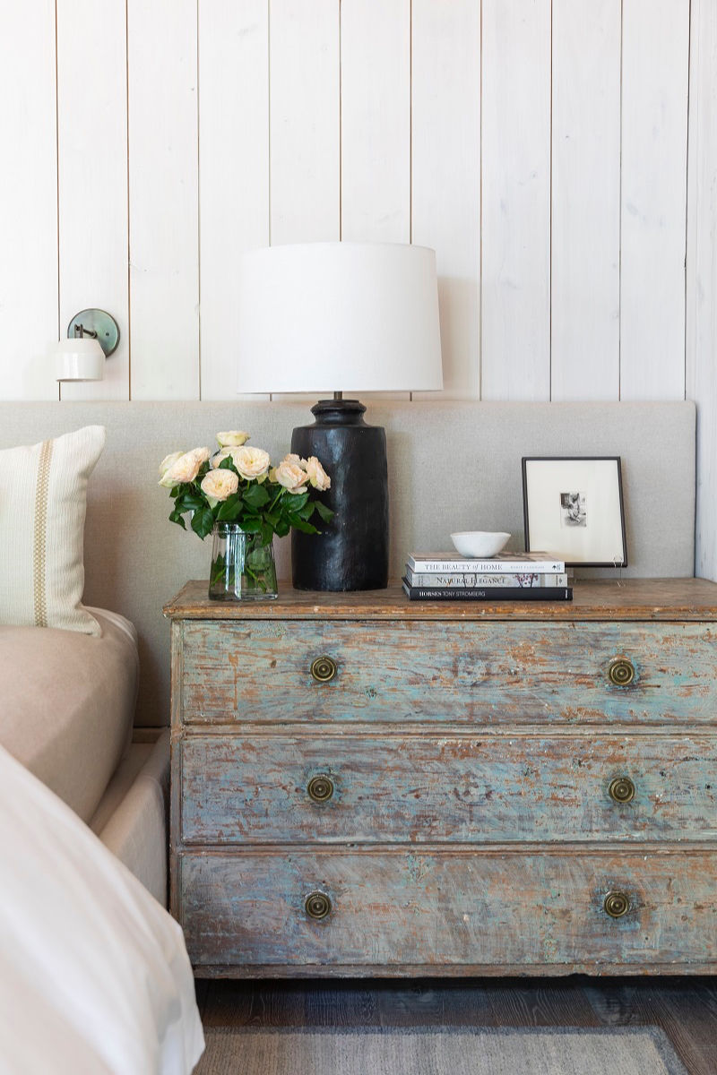 Rustic bedside table with flowers, books, white lamp, and framed photo. Light wood panel wall and cozy bed create a serene mood.