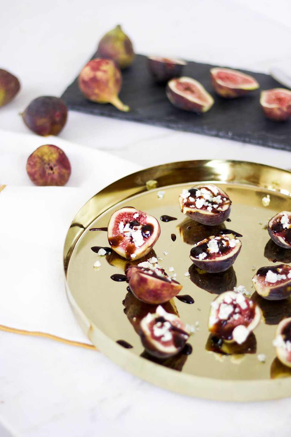 Halved figs with blue cheese and balsamic glaze on a gold tray. Whole and halved figs on a black slate in the background. White surface.
