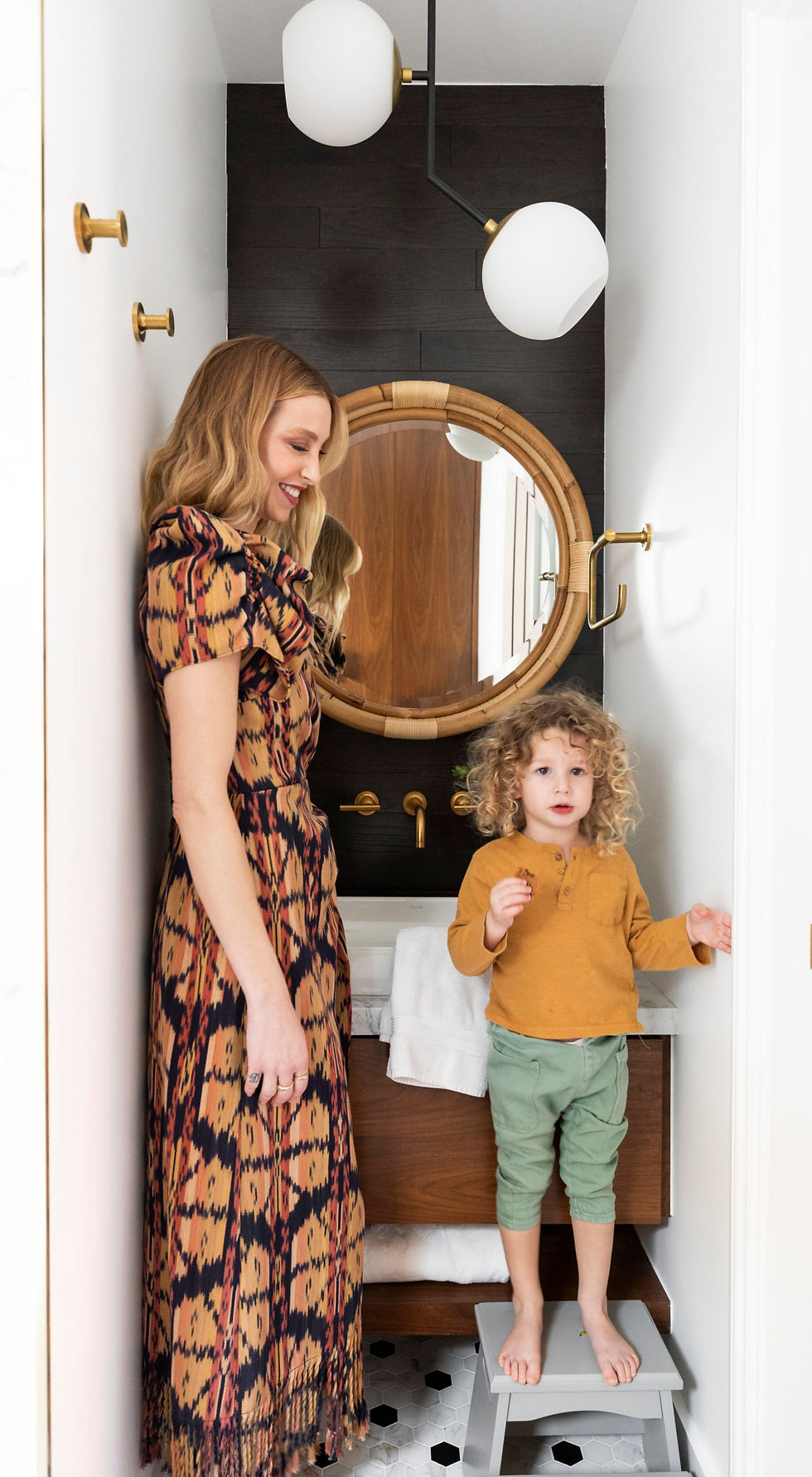 Whitney Port in patterned dress smiles at child in mustard shirt, standing on stool in modern bathroom. Round mirror and black wall accent.