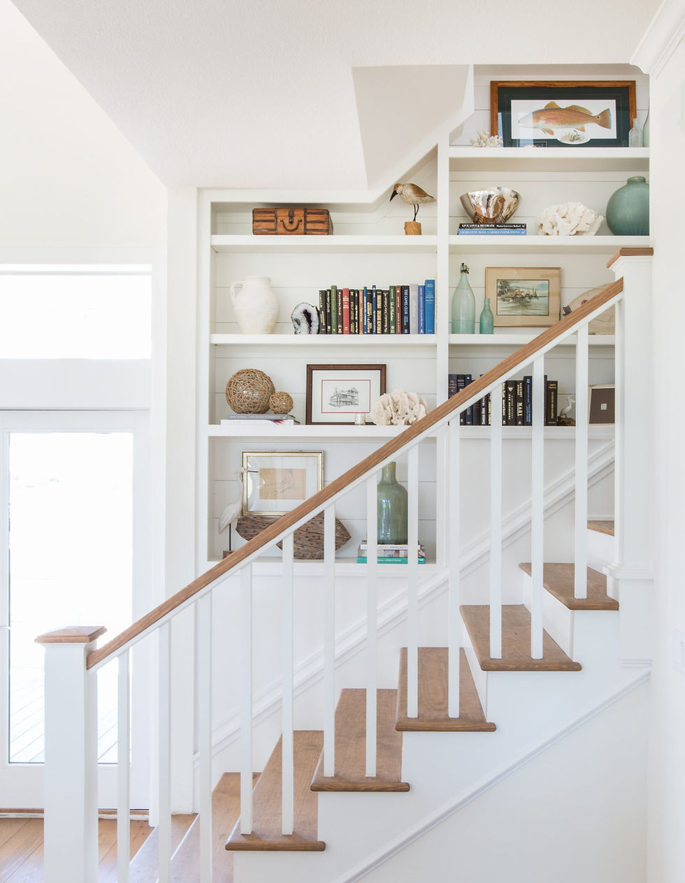 White stairway with wooden steps, bookshelf filled with books, art, and decor items, like vases and coral. Bright, minimalistic room.